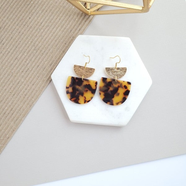 Harper Tortoise Earrings made of dangling tortoiseshell and 18K gold-plated finish are displayed on a hexagonal white marble surface, beautifully complementing the Harper Necklace.