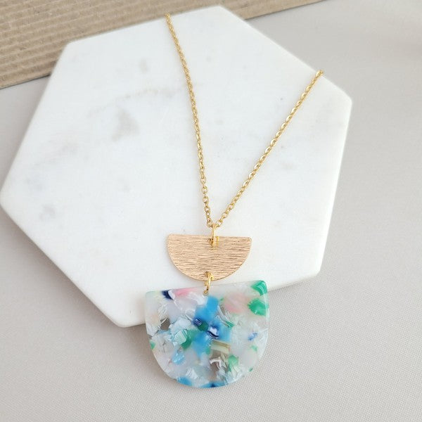 A Harper Spring Fling Necklace with a gold-plated chain and a pendant featuring a semi-circle of acrylic colors and a gold semi-circle shape above it lies on a hexagonal white marble surface.