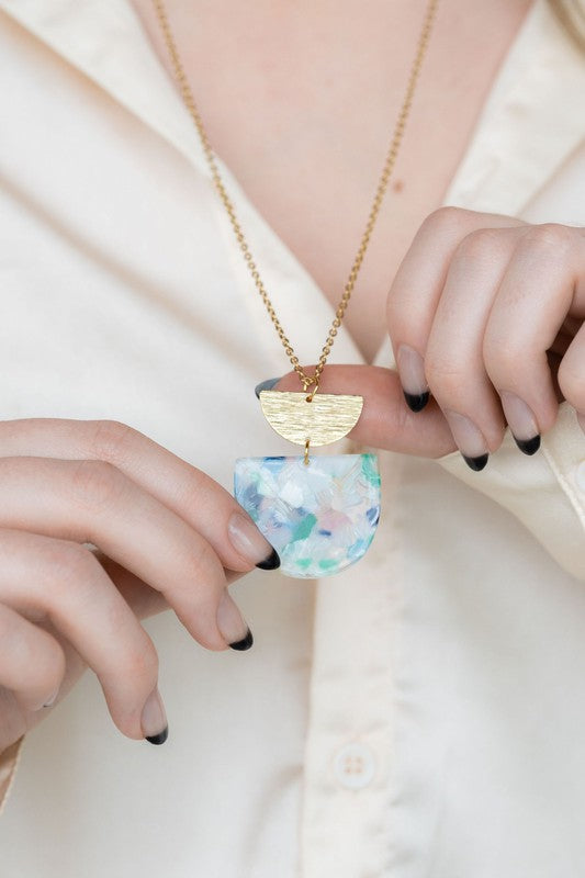 A Harper Spring Fling Necklace with a gold-plated chain and a pendant featuring a semi-circle of acrylic colors and a gold semi-circle shape above it lies on a hexagonal white marble surface.