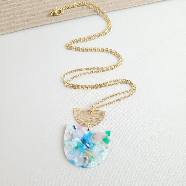 A Harper Spring Fling Necklace with a gold-plated chain and a pendant featuring a semi-circle of acrylic colors and a gold semi-circle shape above it lies on a hexagonal white marble surface.