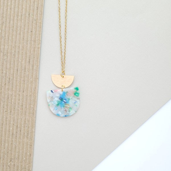 A Harper Spring Fling Necklace with a gold-plated chain and a pendant featuring a semi-circle of acrylic colors and a gold semi-circle shape above it lies on a hexagonal white marble surface.