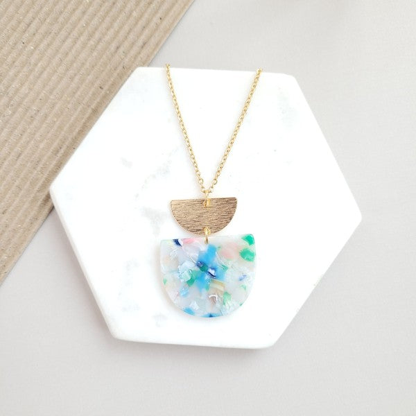 A Harper Spring Fling Necklace with a gold-plated chain and a pendant featuring a semi-circle of acrylic colors and a gold semi-circle shape above it lies on a hexagonal white marble surface.