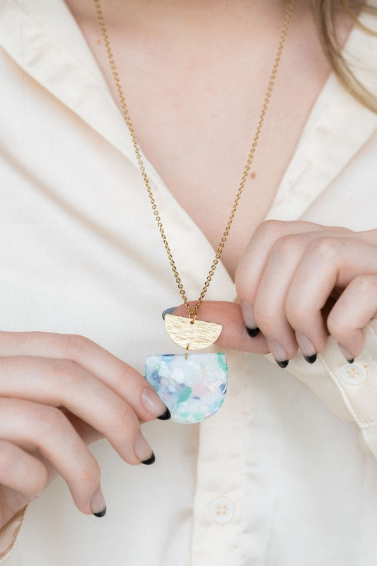 A Harper Spring Fling Necklace with a gold-plated chain and a pendant featuring a semi-circle of acrylic colors and a gold semi-circle shape above it lies on a hexagonal white marble surface.