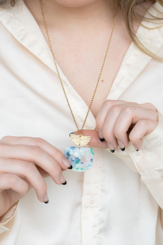 A Harper Spring Fling Necklace with a gold-plated chain and a pendant featuring a semi-circle of acrylic colors and a gold semi-circle shape above it lies on a hexagonal white marble surface.