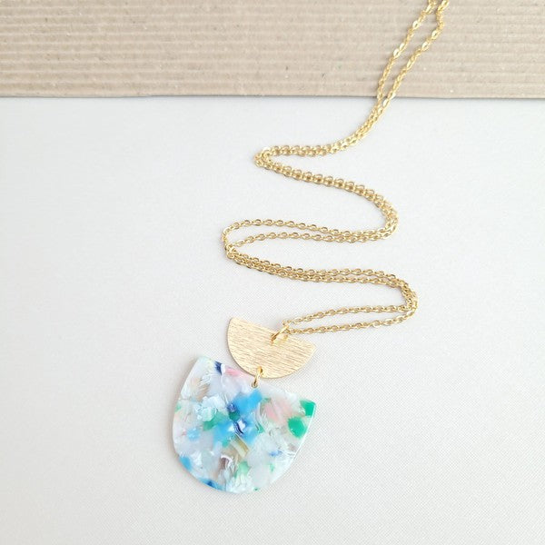 A Harper Spring Fling Necklace with a gold-plated chain and a pendant featuring a semi-circle of acrylic colors and a gold semi-circle shape above it lies on a hexagonal white marble surface.