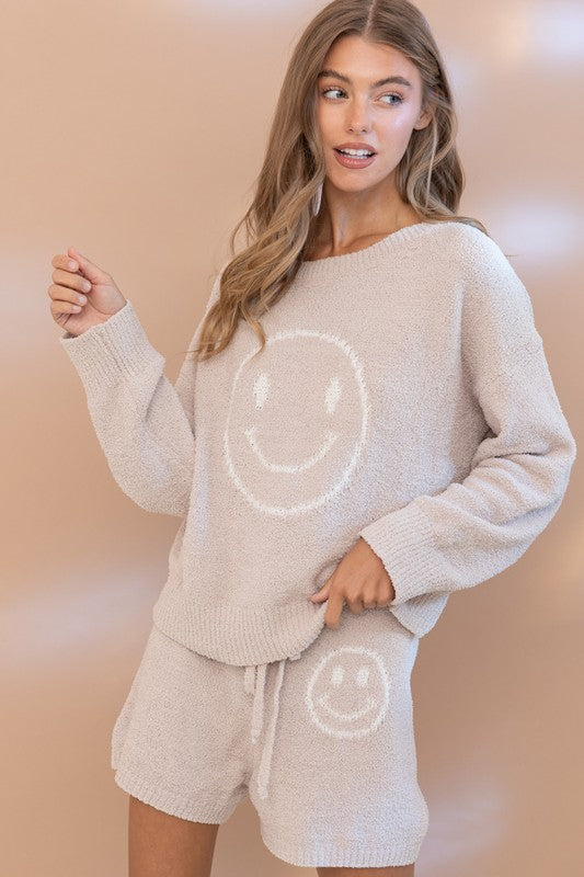 A person is kneeling on a bed, exuding relaxed comfort in the Cozy Soft Top with Shorts Set. The bright pink knit ensemble, adorned with smiley faces, stands out against the backdrop of a wooden wall and a macramé wall hanging.