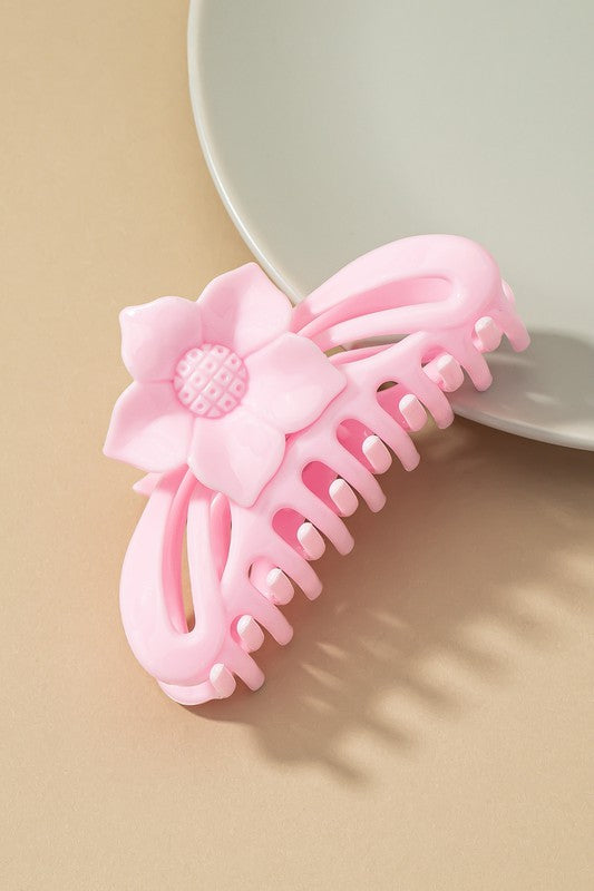 A large hair claw clip featuring a flower handle in a pink hue sits lightly on a beige surface beside a white plate.