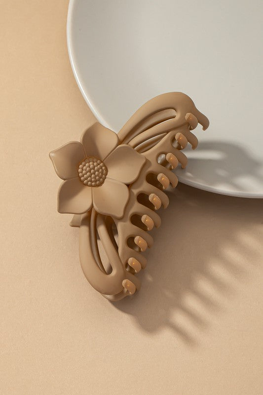 A large hair claw clip featuring a flower handle in a pink hue sits lightly on a beige surface beside a white plate.