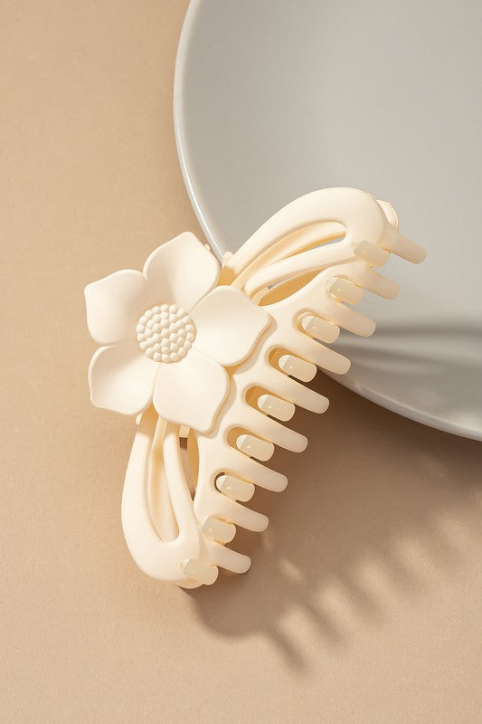 A large hair claw clip featuring a flower handle in a pink hue sits lightly on a beige surface beside a white plate.