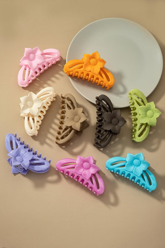 A large hair claw clip featuring a flower handle in a pink hue sits lightly on a beige surface beside a white plate.