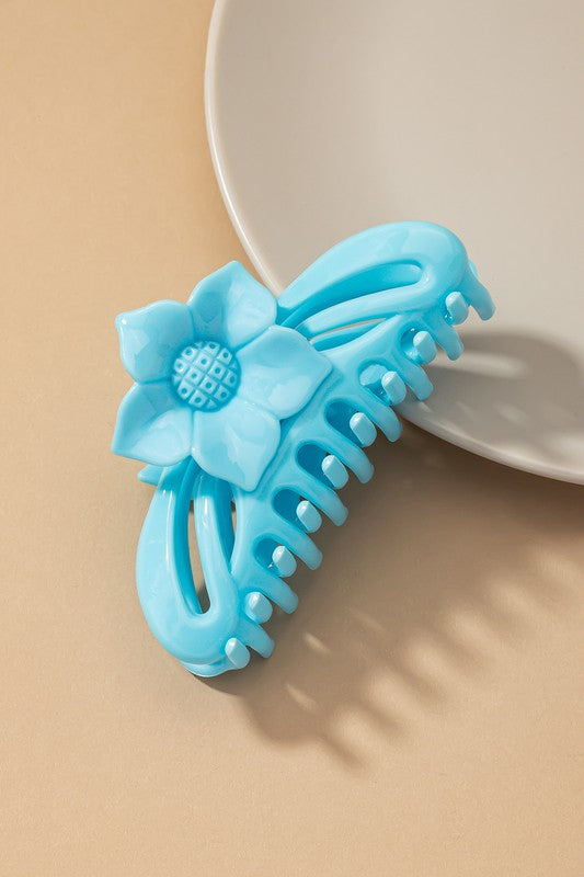 A large hair claw clip featuring a flower handle in a pink hue sits lightly on a beige surface beside a white plate.