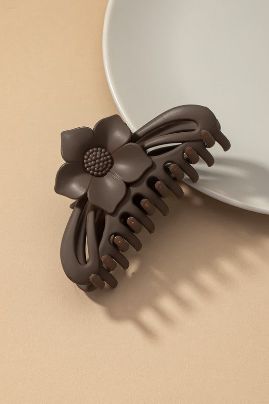 A large hair claw clip featuring a flower handle in a pink hue sits lightly on a beige surface beside a white plate.