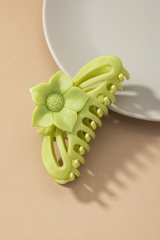 A large hair claw clip featuring a flower handle in a pink hue sits lightly on a beige surface beside a white plate.