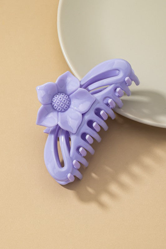 A large hair claw clip featuring a flower handle in a pink hue sits lightly on a beige surface beside a white plate.