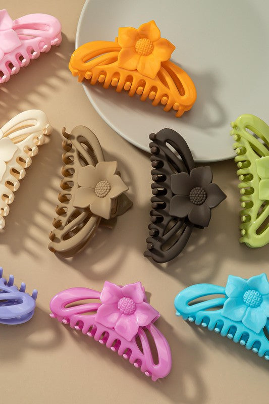 A large hair claw clip featuring a flower handle in a pink hue sits lightly on a beige surface beside a white plate.