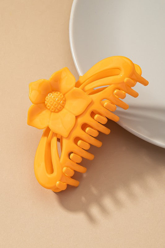 A large hair claw clip featuring a flower handle in a pink hue sits lightly on a beige surface beside a white plate.