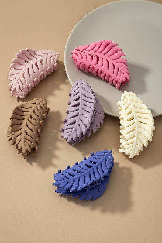 A set of six feather-shaped acrylic hair claw clips in a matte Morandi color palette—pink, beige, purple, brown, blue, and ivory—are stylishly arranged on a grey plate against a brown surface.