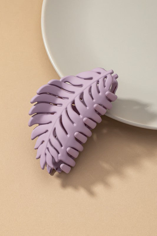 A set of six feather-shaped acrylic hair claw clips in a matte Morandi color palette—pink, beige, purple, brown, blue, and ivory—are stylishly arranged on a grey plate against a brown surface.