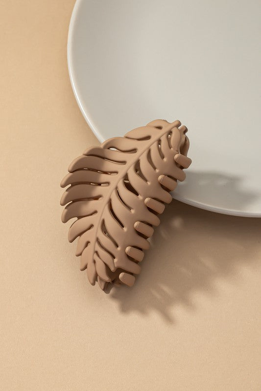 A set of six feather-shaped acrylic hair claw clips in a matte Morandi color palette—pink, beige, purple, brown, blue, and ivory—are stylishly arranged on a grey plate against a brown surface.