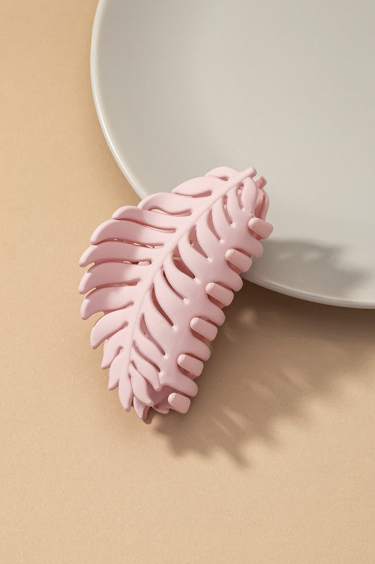 A set of six feather-shaped acrylic hair claw clips in a matte Morandi color palette—pink, beige, purple, brown, blue, and ivory—are stylishly arranged on a grey plate against a brown surface.