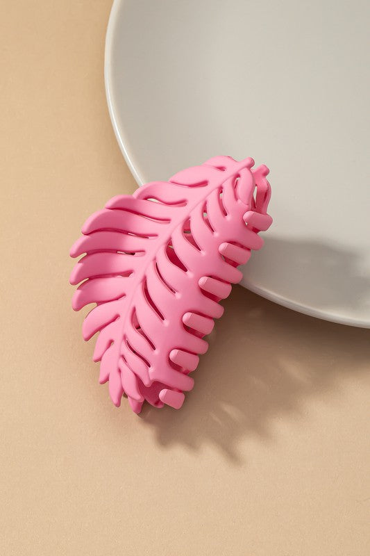 A set of six feather-shaped acrylic hair claw clips in a matte Morandi color palette—pink, beige, purple, brown, blue, and ivory—are stylishly arranged on a grey plate against a brown surface.