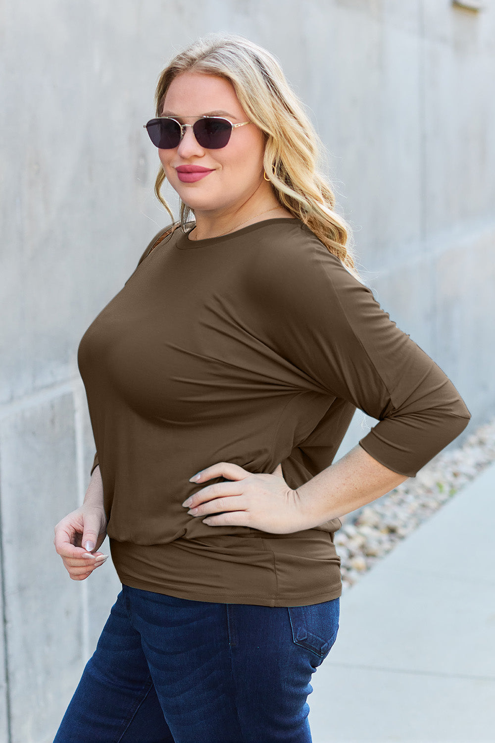 A woman in a slightly stretchy, brown Basic Bae Full Size Round Neck Batwing Sleeve top and blue jeans stands against a concrete wall, holding a brown handbag.