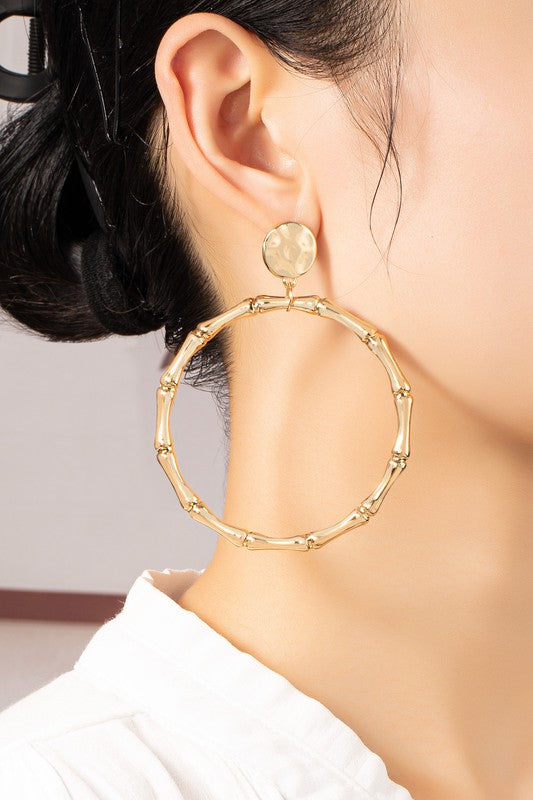 Close-up of a person wearing the Large Statement Bamboo Hoop Earrings, featuring a shiny gold plating and a circular stud, styled in an updo hairstyle, paired with a white top.