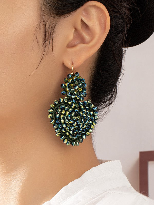 A close-up shot captures a person wearing large, sparkly glass beaded heart drop earrings in vibrant shades of green and blue. The individual has dark hair pulled back and is dressed in a white top.