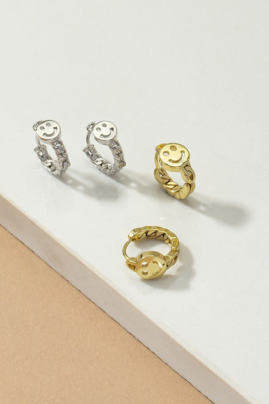 A pair of Smiley face huggie hoop earrings with gleaming gold and silver plating are displayed on a light surface.