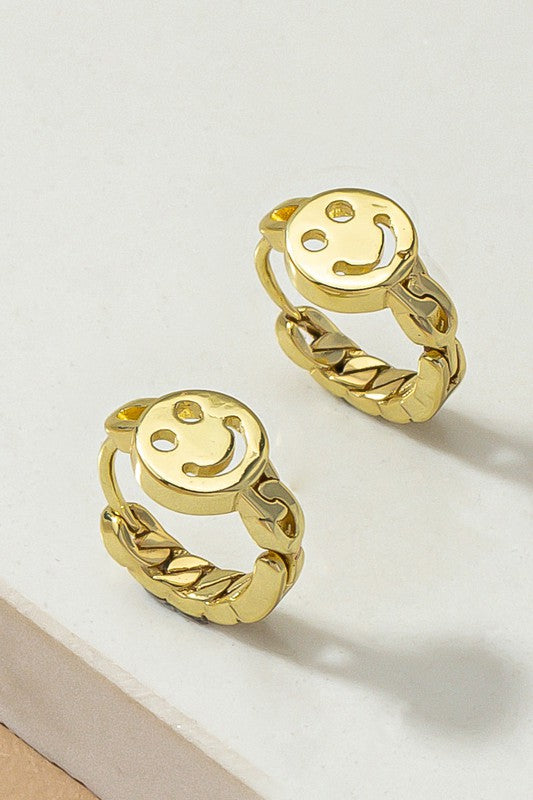 A pair of Smiley face huggie hoop earrings with gleaming gold and silver plating are displayed on a light surface.