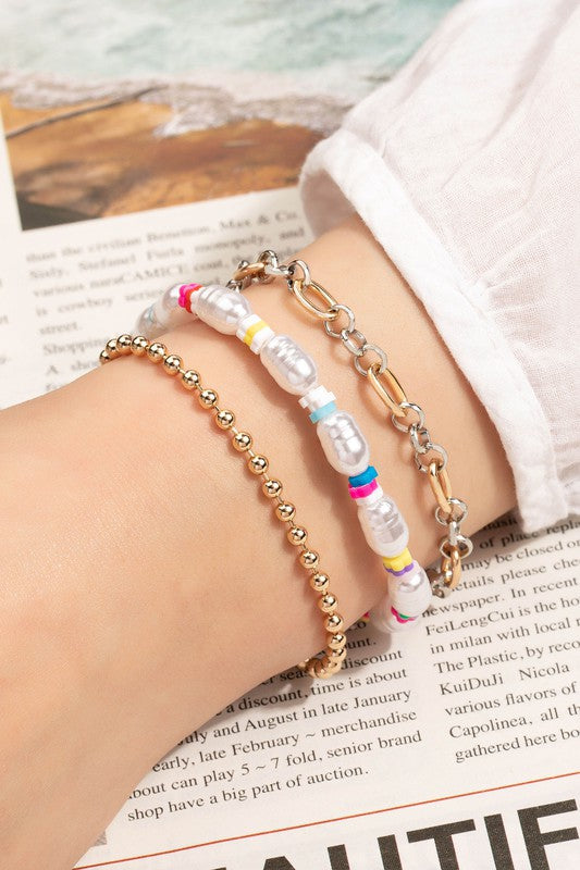 A wrist showcases the exquisite Pearl and Chain Multicolor Bracelets Set, featuring a gold beaded bracelet, one with multicolored beads and faux pearls, and a sleek silver chain link. Behind it, you can see a newspaper and part of a crisp white sleeve.