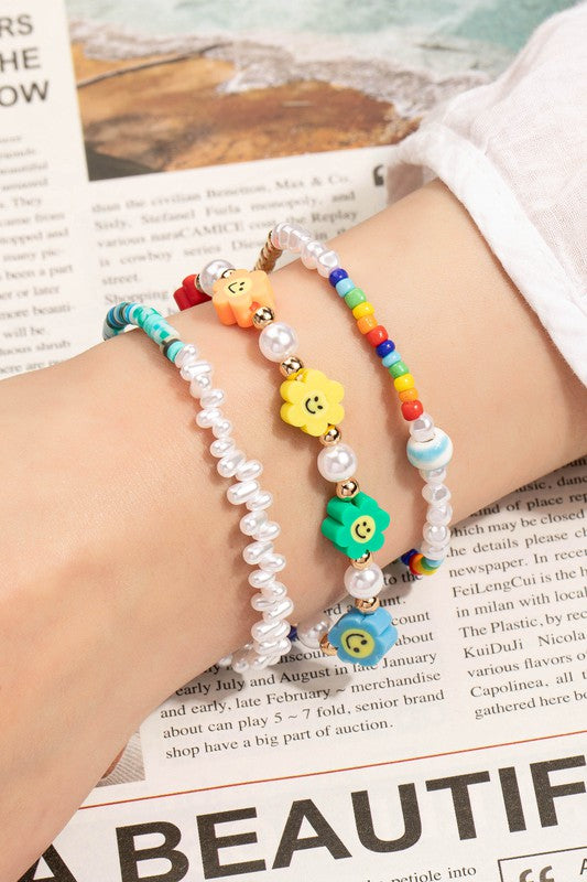 A person wears three vibrant beaded bracelets, including a Smiley face and pearl layered bracelet and one adorned with flower beads, over a page of a newspaper.