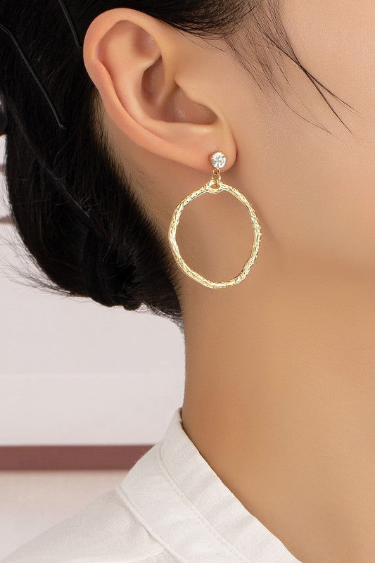 A person wearing the Hammered hoop drop earrings featuring a gold textured design with shiny gold plating, paired with a small rhinestone stud on their ear.