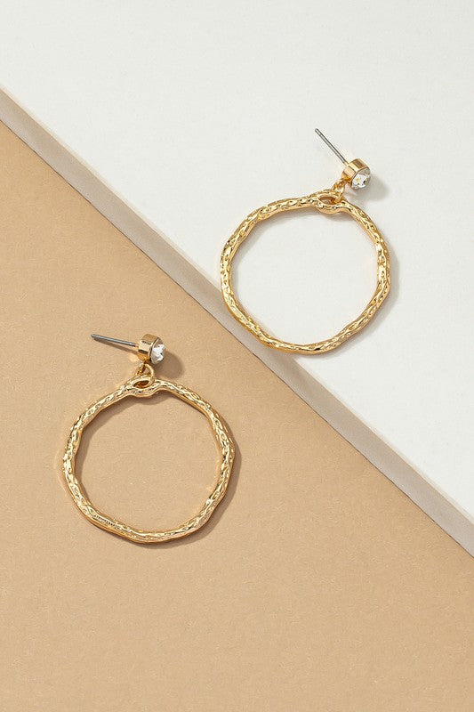 A person wearing the Hammered hoop drop earrings featuring a gold textured design with shiny gold plating, paired with a small rhinestone stud on their ear.