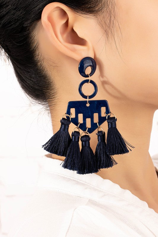 A close-up of someone wearing eye-catching earrings featuring a bold, blue geometric acrylic design with multiple dangling tassels.