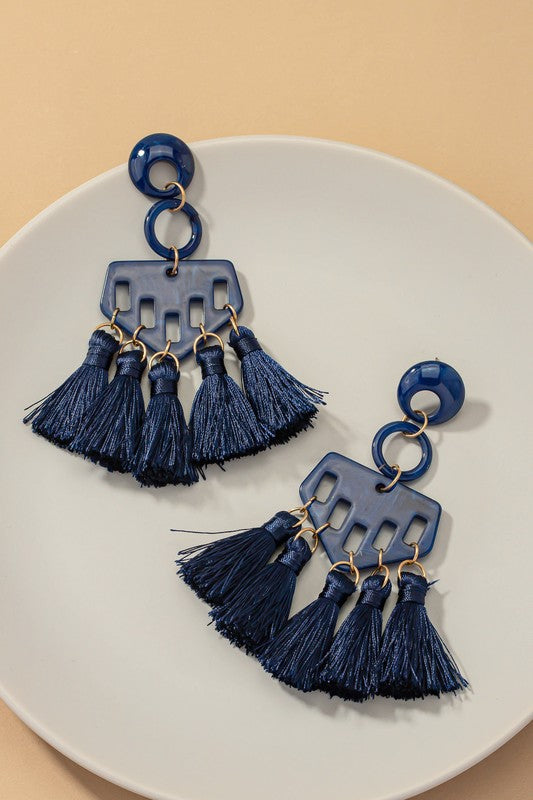 A close-up of someone wearing eye-catching earrings featuring a bold, blue geometric acrylic design with multiple dangling tassels.