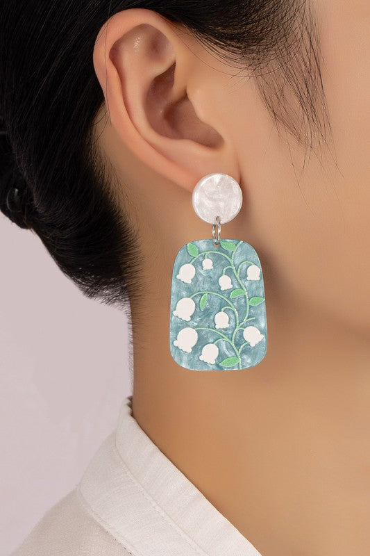 These acetate drop earrings are lightweight and feature a floral design with white flowers and green stems set against a gray background, finished off with a circular top piece.