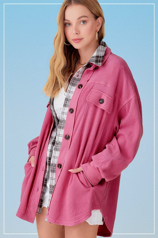 A woman is wearing a pink Sunset Fleece Jacket, buttoned up over a white and black plaid shirt-dress, standing against a blue background. The vibrant colors of her outfit create a striking contrast, emphasizing the cozy and warm outerwear she has chosen.