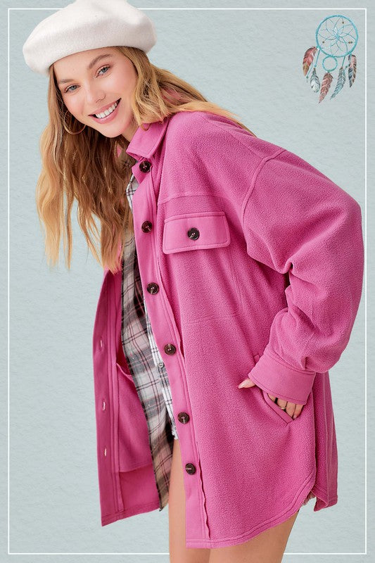 A woman is wearing a pink Sunset Fleece Jacket, buttoned up over a white and black plaid shirt-dress, standing against a blue background. The vibrant colors of her outfit create a striking contrast, emphasizing the cozy and warm outerwear she has chosen.