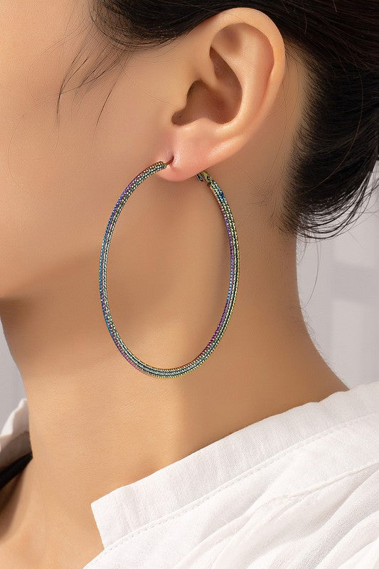 The Holographic Textured Hoop Earrings, featuring a large metallic design with multicolored holographic plating, are set on a white and beige surface.