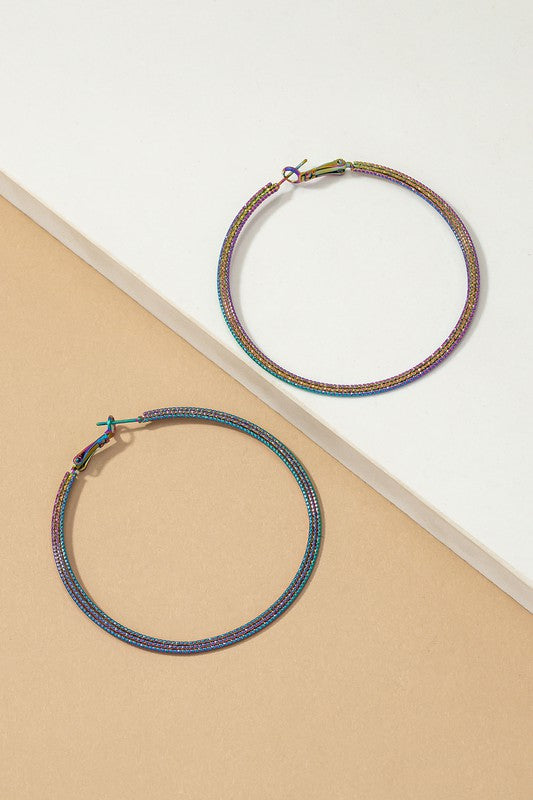 The Holographic Textured Hoop Earrings, featuring a large metallic design with multicolored holographic plating, are set on a white and beige surface.