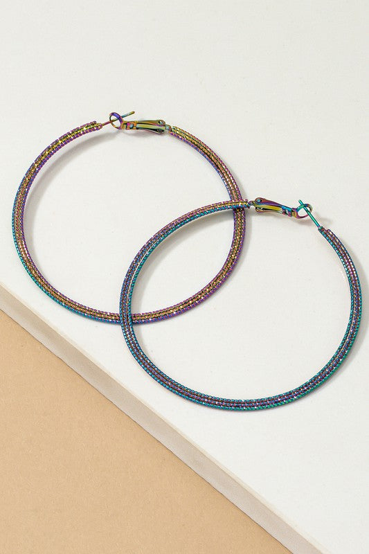 The Holographic Textured Hoop Earrings, featuring a large metallic design with multicolored holographic plating, are set on a white and beige surface.