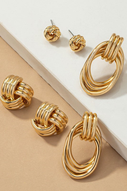 The Premium Trio Metal Knot and Hoop Earrings, displayed on a marble-patterned card, showcase three pairs of unique woven knot designs all embellished with shiny gold plating against a tan fabric backdrop.