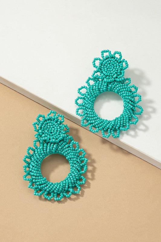 A pair of handmade statement seed bead earrings, featuring intricate circular designs in green, is beautifully showcased against a two-toned beige and white surface.