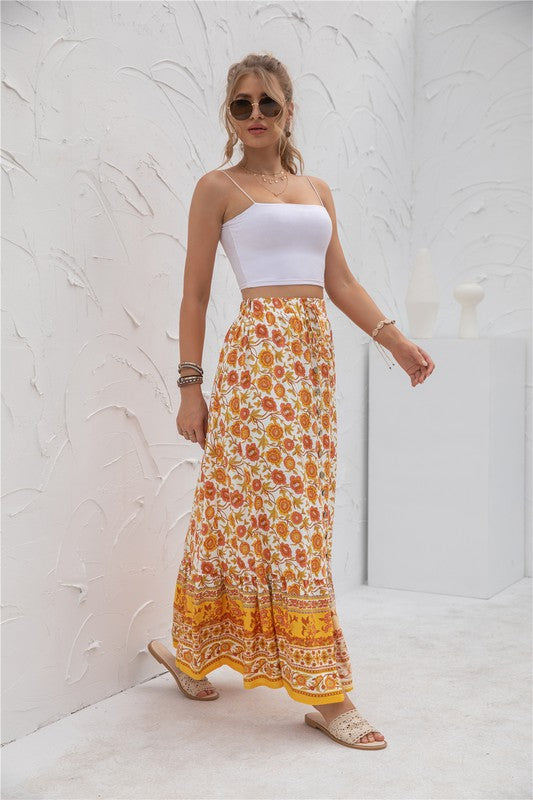 A woman stands against a light-textured wall wearing a white crop top, sunglasses, and holding a woven hat. She complements her look with the Womens Print Maxi Skirt, an eye-catching long red skirt with patterns.