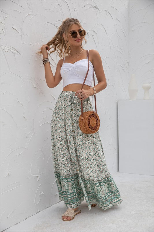 A woman stands against a light-textured wall wearing a white crop top, sunglasses, and holding a woven hat. She complements her look with the Womens Print Maxi Skirt, an eye-catching long red skirt with patterns.