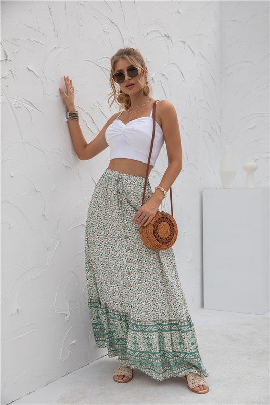 A woman stands against a light-textured wall wearing a white crop top, sunglasses, and holding a woven hat. She complements her look with the Womens Print Maxi Skirt, an eye-catching long red skirt with patterns.