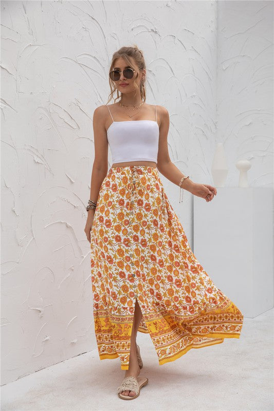 A woman stands against a light-textured wall wearing a white crop top, sunglasses, and holding a woven hat. She complements her look with the Womens Print Maxi Skirt, an eye-catching long red skirt with patterns.