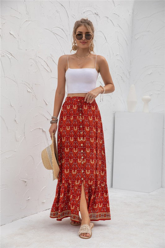 A woman stands against a light-textured wall wearing a white crop top, sunglasses, and holding a woven hat. She complements her look with the Womens Print Maxi Skirt, an eye-catching long red skirt with patterns.