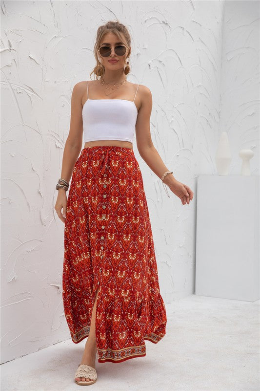 A woman stands against a light-textured wall wearing a white crop top, sunglasses, and holding a woven hat. She complements her look with the Womens Print Maxi Skirt, an eye-catching long red skirt with patterns.